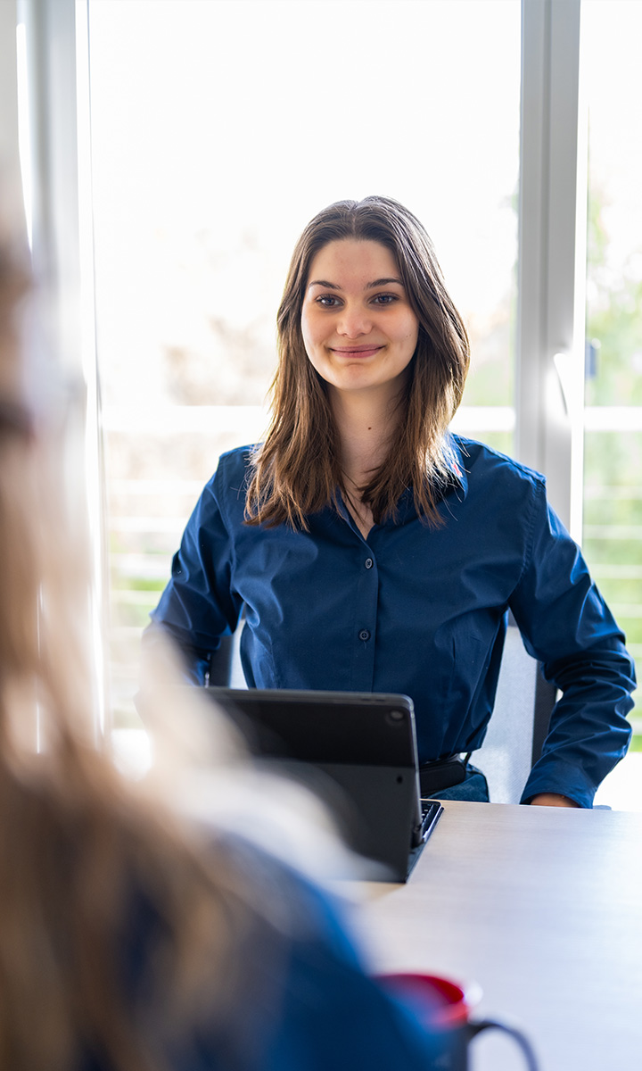 Eine junge Frau in blauer Bluse sitzt lächelnd an einem Schreibtisch mit Tablet gegenüber einer weiteren Person.