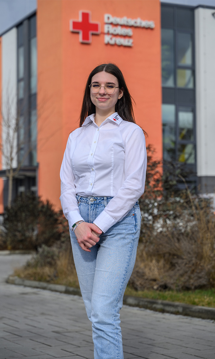 Eine junge Frau mit Brille, weißer Bluse und Jeans steht lächelnd vor einem Gebäude mit dem Logo des Deutschen Roten Kreuzes.