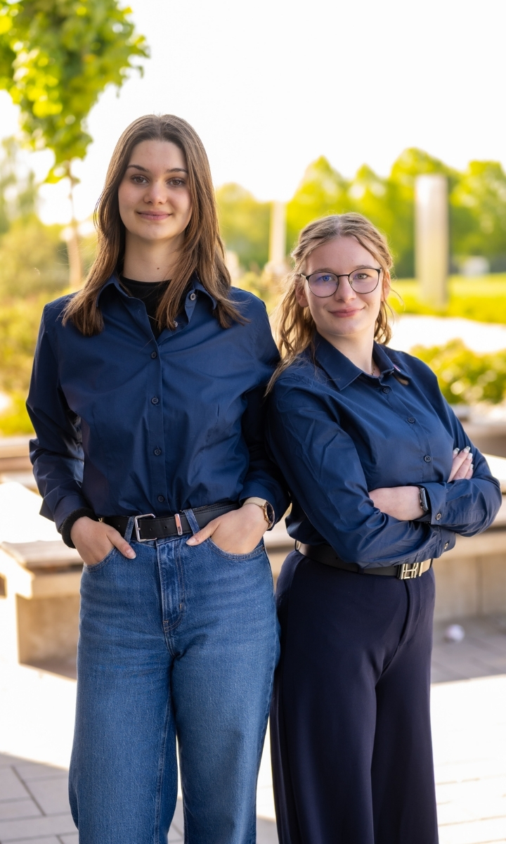 Zwei Auszubildende aus der DRK-Verwaltungs-Ausbildung stehen lachend vor dem Eingang des Verwaltungsgebäudes.