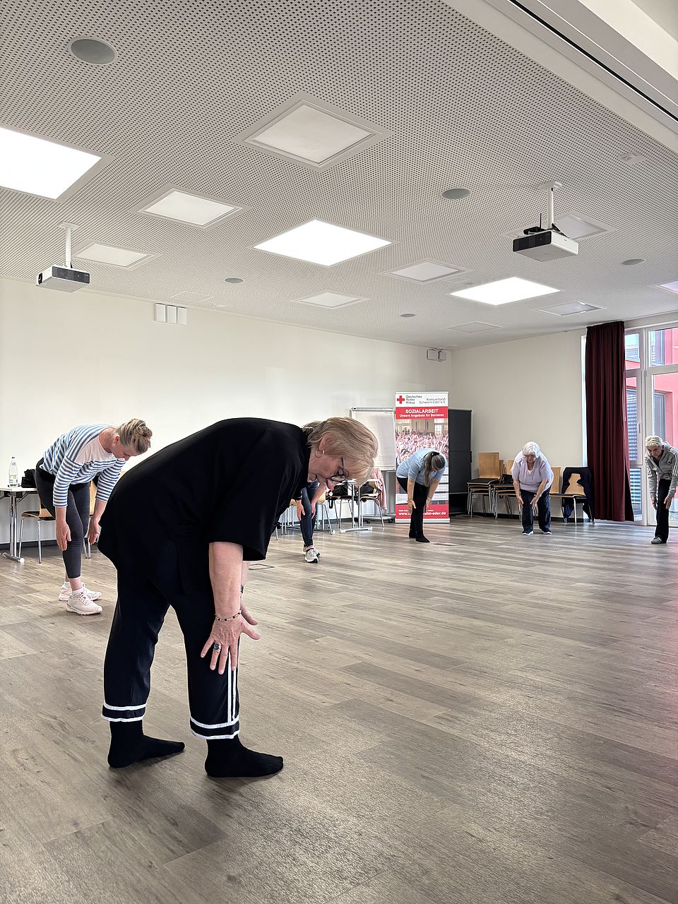 Gruppe älterer Frauen beim Qigong-Training in einem lichtdurchfluteten Raum, angeleitet von einer Trainerin in schwarzer Kleidung.