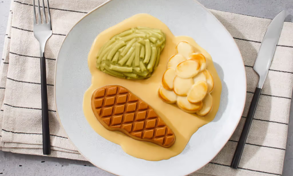 Ein weißer Teller, von oben fotografiert, mit einer pürierten und optisch ansprechend in Form gebrachten Mahlzeit (passierte Kost). Auf einem Spiegel aus glatter, heller Soße liegen ein geformtes Stück Fleisch mit nachgebildetem Grillmuster, eine Portion grüne Bohnen und geschichtete Kartoffelscheiben. Alle Komponenten weisen eine homogene, sehr weiche Konsistenz auf. Der Teller steht auf einem beigen Tischset mit feinen dunklen Streifen. Links neben dem Teller liegt eine Gabel, rechts ein Messer mit schwarzem Griff.