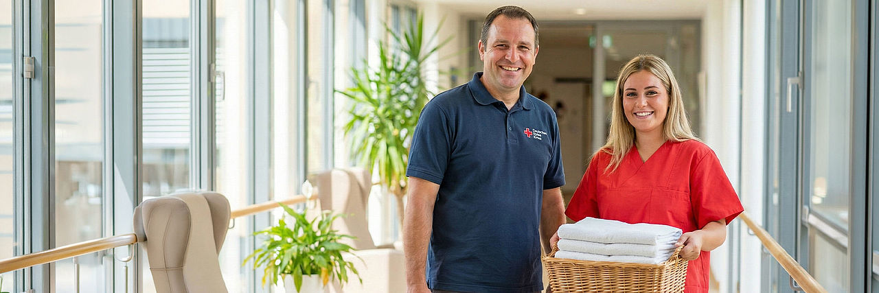 Hausmeister und Pflegekraft des DRK stehen gemeinsam in einem Flur einer Pflegeeinrichtung, die Pflegekraft trägt frische Handtücher, der Hausmeister eine Werkzeugtasche.