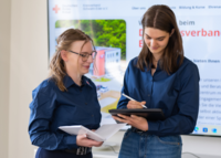 Zwei DRK-Mitarbeiterinnen besprechen sich vor einem Bildschirm mit Notizbuch und Tablet.