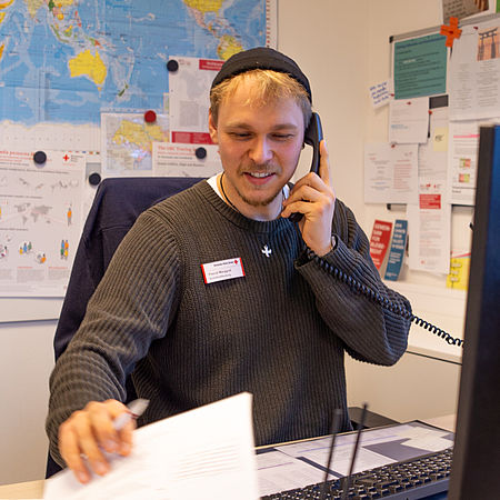 Am Telefon_Pascal_01 Pascal Marggraf, Berater des DRK-Suchdienstes, telefoniert an seinem Schreibtisch und hält dabei ein Dokument in der Hand. Im Hintergrund hängt eine große politische Weltkarte des DRK-Suchdienstes an der Wand.