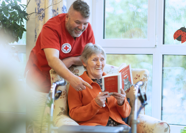 Pflegekraft des DRK lacht gemeinsam mit einer älteren Frau, die in einem Sessel sitzt und ein Buch liest. Beide wirken fröhlich und verbunden. Im Vordergrund ist ein Rollator zu sehen.