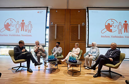 Podiumsdiskussion beim DRK: Fünf Personen aus verschiedenen Generationen und Hintergründen sprechen vor dem Logo des DRK-Suchdienstes mit dem Motto „Suchen. Verbinden. Vereinen.“