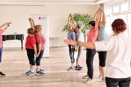 Seniorengruppe bei Gymnastikübung in einem hellen Raum, Arme nach oben gestreckt, im Hintergrund ein Banner des Deutschen Roten Kreuzes.