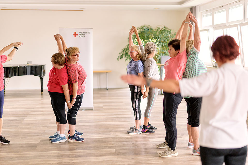 Highres_DRK_Hessen_2728 Seniorengruppe bei Gymnastikübung in einem hellen Raum, Arme nach oben gestreckt, im Hintergrund ein Banner des Deutschen Roten Kreuzes.