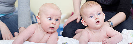 Zwei Babys liegen nebeneinander auf dem Bauch auf einer weißen Decke, schauen aufmerksam nach vorn. Im Hintergrund sind unscharf zwei Erwachsene zu sehen, die neben den Kindern sitzen.“