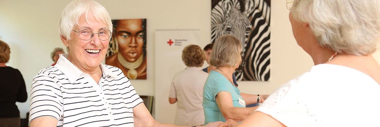 Zwei Seniorinnen tanzen lachend miteinander in einem Gruppenraum
