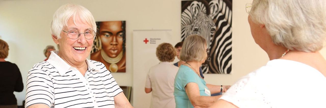 Zwei Seniorinnen tanzen lachend miteinander in einem Gruppenraum
