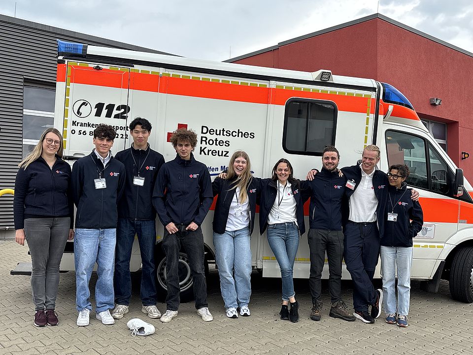 Eine Gruppe von neun jungen DRK-Mitarbeiterinnen und -Mitarbeitern steht lächelnd nebeneinander vor einem Rettungswagen des Deutschen Roten Kreuzes. Alle tragen dunkle DRK-Jacken und Namensschilder, im Hintergrund ist ein modernes Gebäude mit roter Fassade zu sehen.