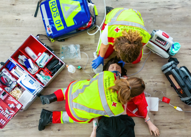 Rettungssanitäter und Notfallsanitäter bei der Arbeit an einem Patienten. Das Equipment liegt um das Rettungsdienstpersonla herum. Zu sehen ist ein Notfallkoffer , Beatmungsrucksack, EKG, Absaugpumpe und viele Materialien wie Verbände und Medikamenteampullen