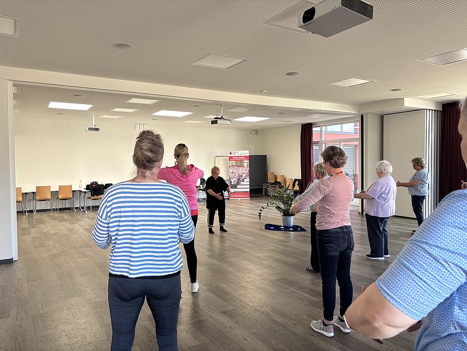 Gruppe älterer Frauen beim Qigong-Training in einem lichtdurchfluteten Raum, angeleitet von einer Trainerin in schwarzer Kleidung.