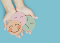 Zwei Hände halten drei bunte Smiley-Gesichter mit unterschiedlichen Emotionen vor hellblauem Hintergrund.