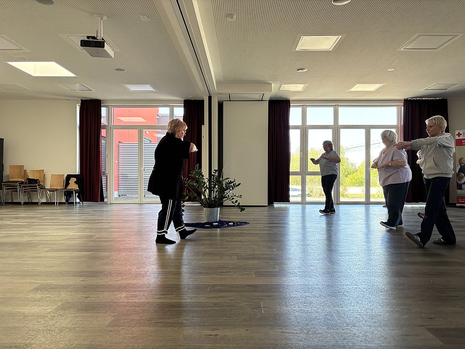 Gruppe älterer Frauen beim Qigong-Training in einem lichtdurchfluteten Raum, angeleitet von einer Trainerin in schwarzer Kleidung.