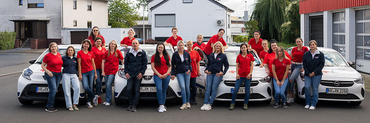 Teamfoto des Teams der ambulanten Pflege des DRK Schwalm-Eder in roten Tshirts zwischen dem Fuhrpark