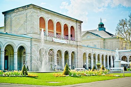 Arkadenbau in Bad Kissingen mit Springbrunnen, Blumenbeeten und gepflegter Grünanlage bei sonnigem Wetter.