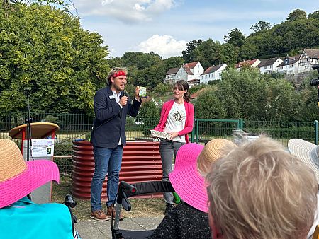 Ein Mann mit rotem Stirnband und Mikrofon spricht bei einer Outdoor-Veranstaltung vor älteren Menschen mit bunten Hüten. Neben ihm steht eine Frau mit Eierkarton und lächelt. Im Hintergrund sind grüne Bäume, Fachwerkhäuser und ein Gartenbeet zu sehen.