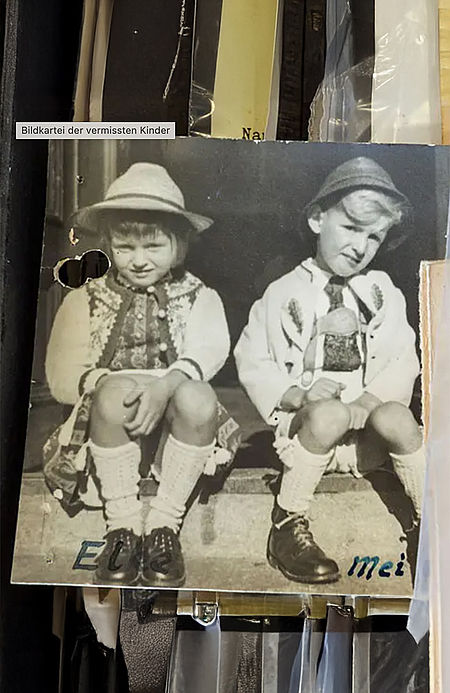 Schwarz-weiß-Foto aus der Bildkartei vermisster Kinder: Zwei kleine Kinder in traditioneller Tracht sitzen nebeneinander auf einer Treppe. Die Karte zeigt Gebrauchsspuren und ist handbeschriftet mit den Namen „Els“ und „Mei“.