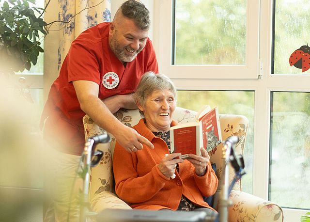 Pflegekraft im DRK-Shirt liest gemeinsam mit einer Seniorin in einem Buch
