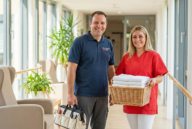 Hausmeister und Pflegekraft des DRK stehen gemeinsam in einem Flur einer Pflegeeinrichtung, die Pflegekraft trägt frische Handtücher, der Hausmeister eine Werkzeugtasche.