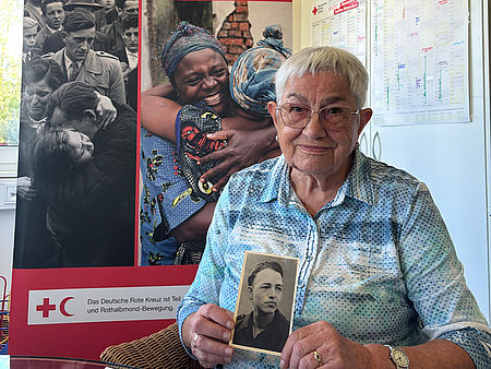 Ältere Frau hält ein historisches Porträtfoto eines jungen Soldaten in den Händen, im Hintergrund ein Roll-up des DRK-Suchdienstes „Restoring Family Links".