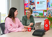 Zwei Frauen im Gespräch an einem Bürotisch mit Tablet und roten Tassen des Deutschen Roten Kreuzes.