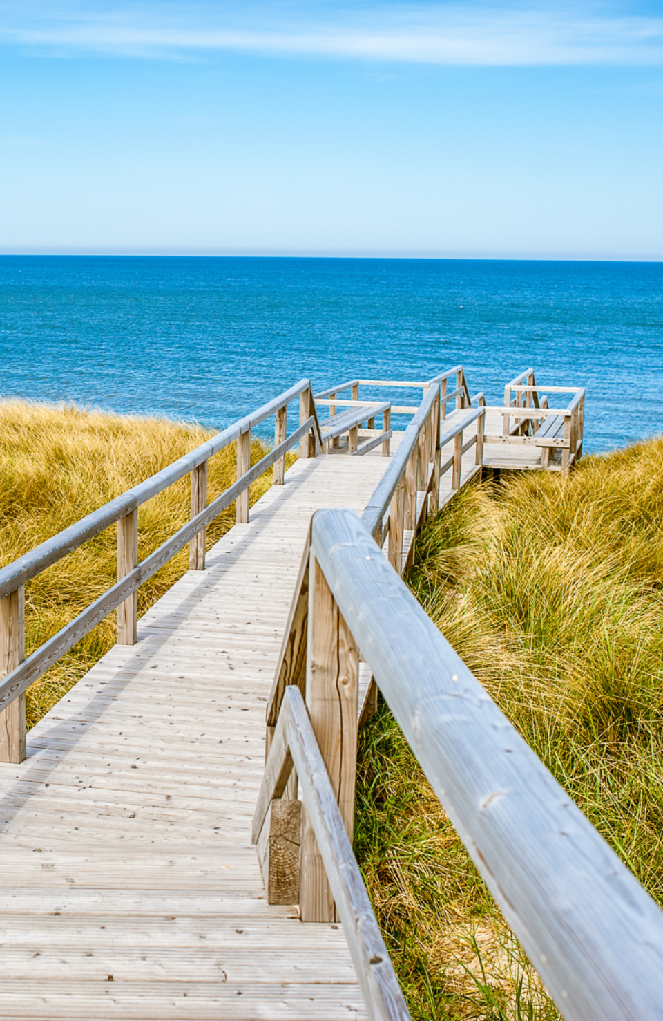 Holzsteg führt durch Dünen zum Meer auf Sylt – strahlend blauer Himmel und ruhige See im Hintergrund.
