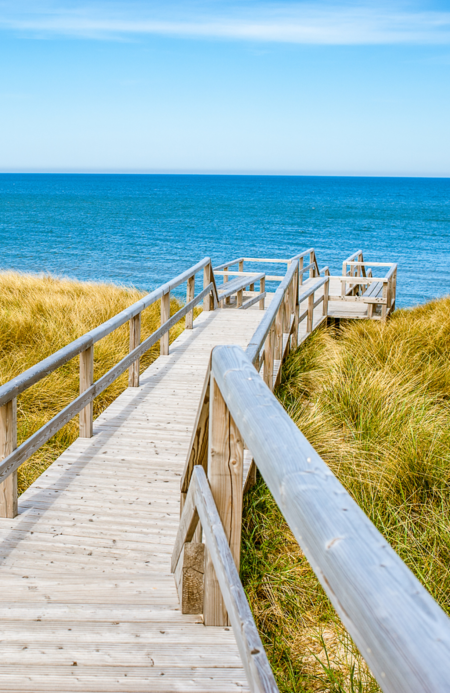 Holzsteg führt durch Dünen zum Meer auf Sylt – strahlend blauer Himmel und ruhige See im Hintergrund.