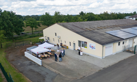 Luftaufnahme des neuen DRK-Rettungsdienststandortes in Wabern-Harle während der Einweihungsfeier am 1. Juni 2026. Vor dem hellen, länglichen Gebäude mit Solarpaneelen auf dem Dach haben sich Personen auf dem gepflasterten Vorplatz versammelt. Einige Gäste sitzen an Bierzeltgarnituren unter zwei weißen DRK-Pavillons, andere stehen in kleinen Gruppen zusammen und unterhalten sich. An der Fassade prangen das Logo des Deutschen Roten Kreuzes sowie ein Schild mit der Notrufnummer 112. Im Hintergrund erstreckt sich eine grüne Landschaft mit Bäumen, Wiesen und einer Landstraße unter einem leicht bewölkten Himmel.