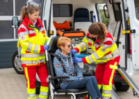 Zwei Rettungssanitäterinnen des DRK versorgen eine Person auf einem Tragestuhl vor dem Rettungswagen.