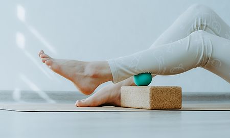 Beine in weißer Sportkleidung bei einer Faszienübung mit einem Massageball auf einem Yogablock.