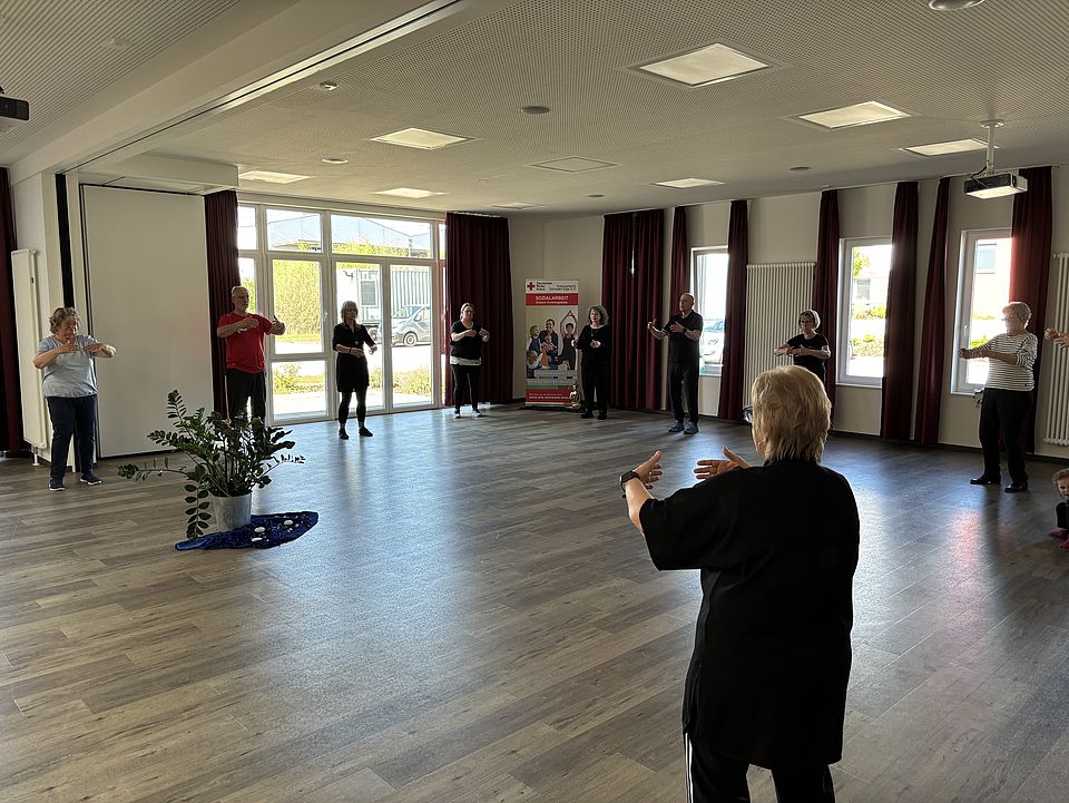 Gruppe älterer Frauen beim Qigong-Training in einem lichtdurchfluteten Raum, angeleitet von einer Trainerin in schwarzer Kleidung.
