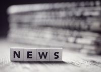 Symbolfoto mit Holzbuchstaben „NEWS“ vor einem Zeitungsstapel, schwarz-weiß gehalten.