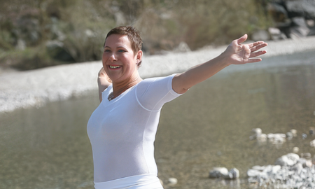 Eine lächelnde Frau mit kurzen Haaren steht mit weit ausgebreiteten Armen im Freien vor einem flachen, steinigen Flussbett.