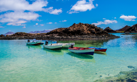 Kleine bunte Boote liegen in ruhigem, türkisfarbenem Wasser vor einer felsigen Küste auf Fuerteventura, im Hintergrund ein dunkler Vulkanküstenstreifen und ein strahlend blauer Himmel mit einzelnen weißen Wolken.