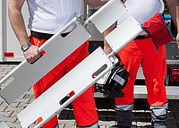 Zwei DRK-Einsatzkräfte in roter Einsatzkleidung tragen eine Trage und medizinisches Equipment vor einem Einsatzfahrzeug.