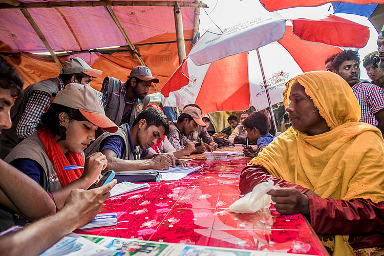 Eine Gruppe von humanitären Helfern in grauen Westen und Kappen sitzt an einem langen, mit einer roten gemusterten Tischdecke bedeckten Tisch im Freien und registriert Menschen unter einem großen rot-weißen Sonnenschirm. Eine Frau in einem leuchtend gelben Hidschab sitzt den Helfern gegenüber, während diese konzentriert Dokumente ausfüllen.